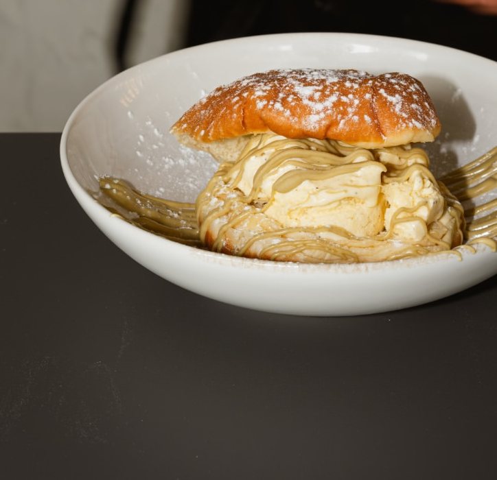 Bola de Berlim polvilhada com açúcar, com gelado e um molho cor de pistáchio por cima, numa taça branca sobre uma mesa escura