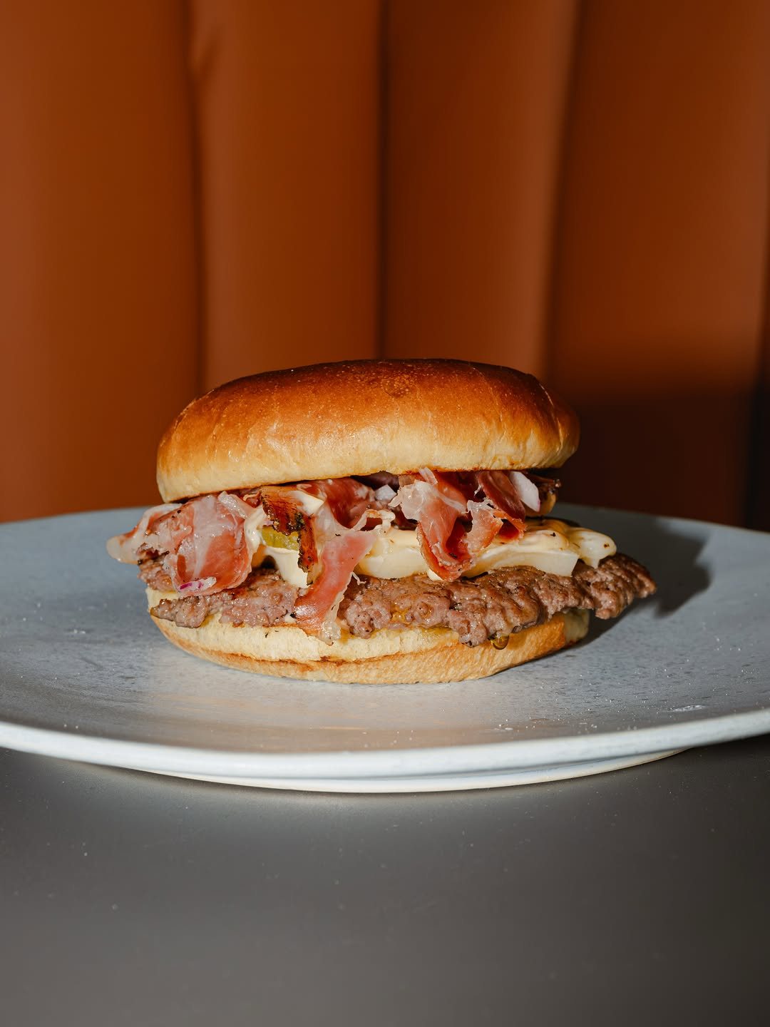 Hambúrguer de carne com fatias finas curadas, queijo e cebola roxa num pão brioche, num prato cinzento, com um fundo cor de laranja